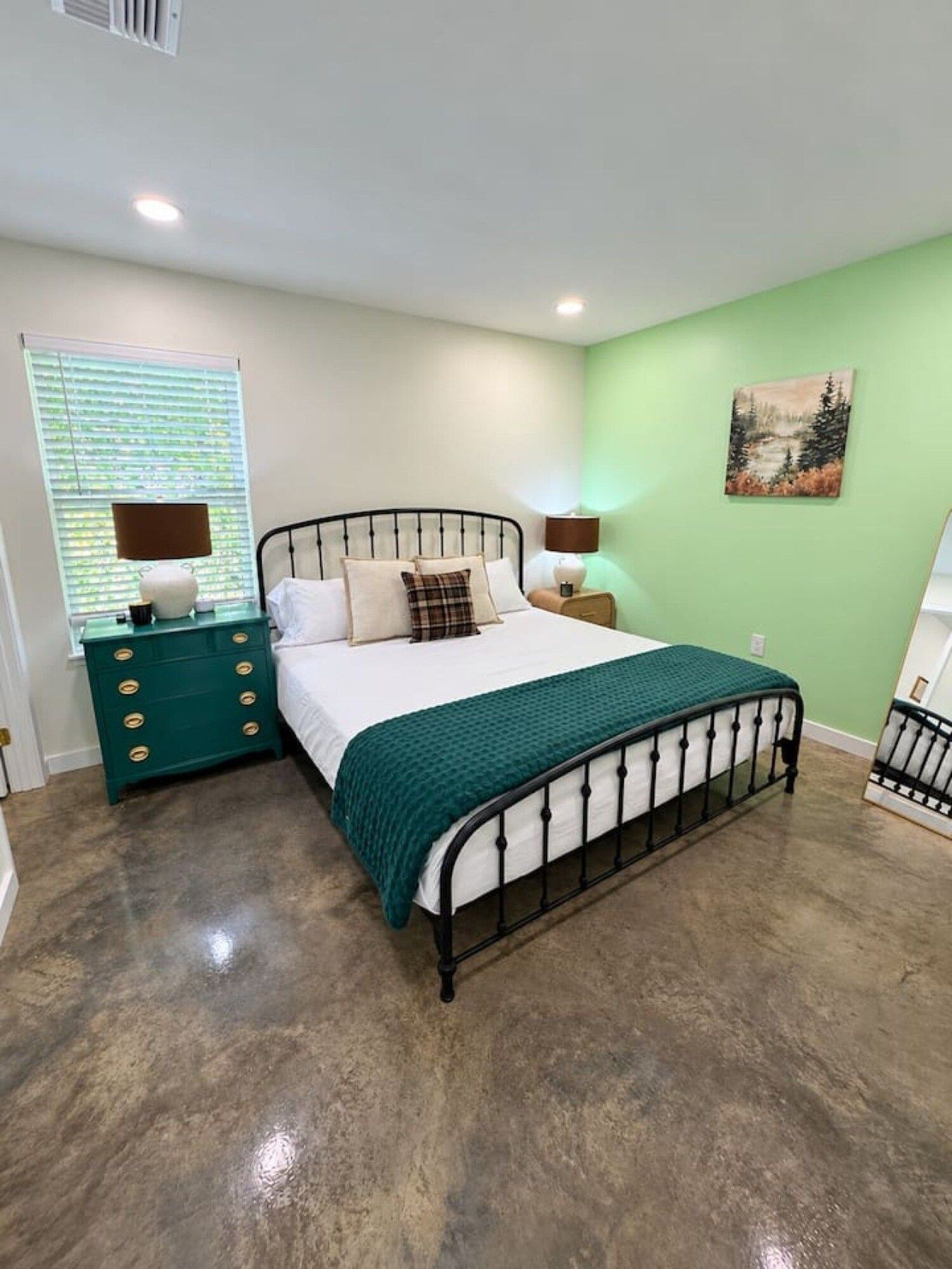 A bedroom with a metal bed frame, a green dresser, a window with blinds, and a green wall with a painting.