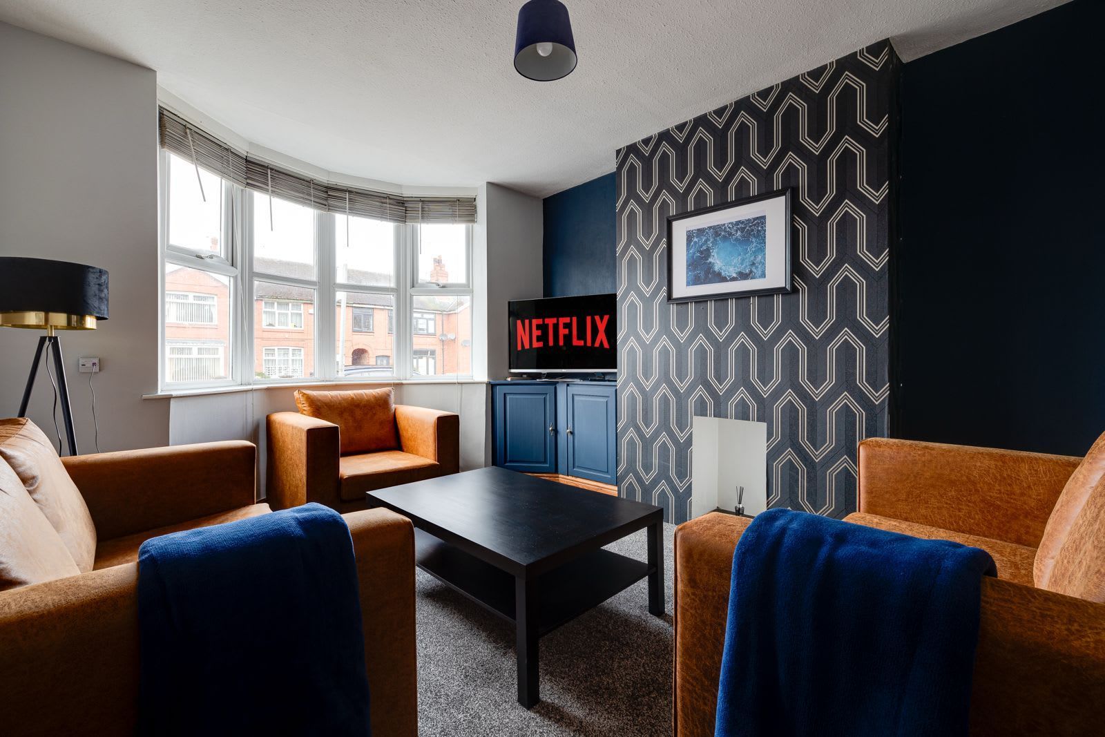 A modern living room with a sofa, armchair, and a coffee table. There is a television on a stand with a Netflix sign, a patterned wall, and a window with blinds.