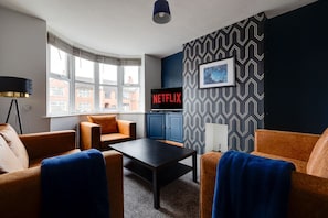 A modern living room with a sofa, armchair, and a coffee table. There is a television on a stand with a Netflix sign, a patterned wall, and a window with blinds. - Bright 3-bed Home Crewe Garden Netflix Wi-fi (Crewe)