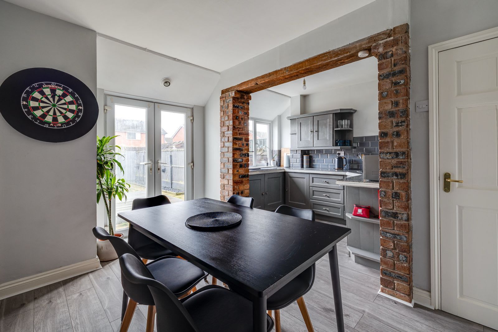 A dining area with a black table and chairs, a dartboard, and a brick archway leading to a kitchen.