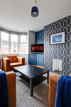A modern living room with a black coffee table, orange armchairs, a blue cabinet, and a wall-mounted TV displaying 'NETFLIX'. - Bright 3-bed Home Crewe Garden Netflix Wi-fi (Crewe)
