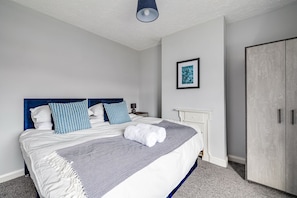A neatly made bed with a blue headboard, a bedside table with a lamp, a framed picture on the wall, and a wardrobe. - Bright 3-bed Home Crewe Garden Netflix Wi-fi (Crewe)