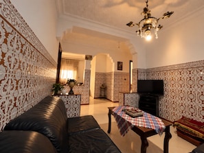A living room with a sofa, a television, a table with a tablecloth, and patterned walls.