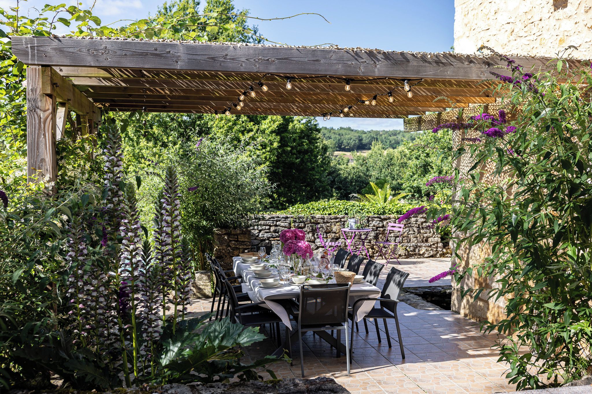 An outdoor dining area with a pergola, stone wall, and lush greenery.