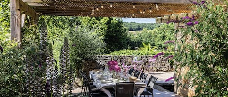 Un área de comedor al aire libre con pérgola, pared de piedra y abundante vegetación.