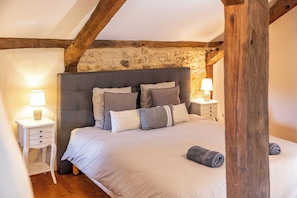 A bedroom with a stone wall, a wooden ceiling beam, a bed with multiple pillows, two bedside tables with lamps, and a rolled towel on the bed. - New Listing (Milhac-de-Nontron)