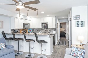 A modern kitchen with white cabinets, a central island with bar stools, a ceiling fan, and a view into the living area. - Bayside 1BR Condo with Pool (Ocean City)