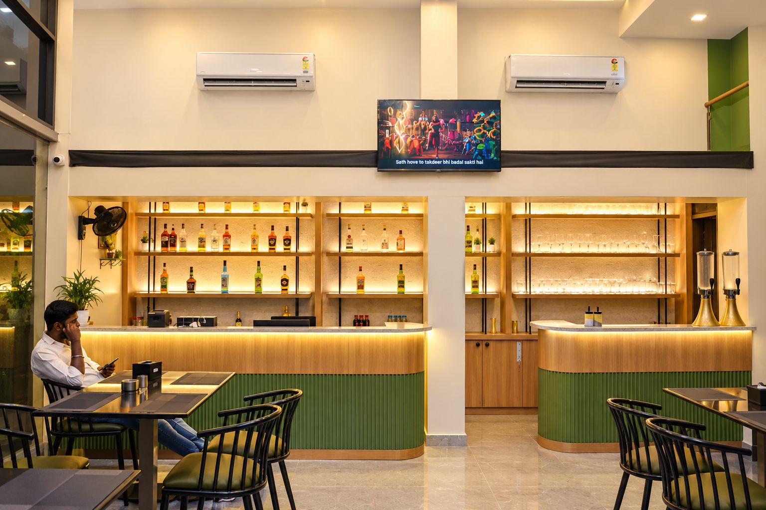 A bar area with shelves of liquor bottles, a TV mounted on the wall, and a person seated at a table.