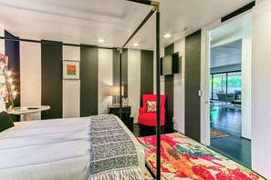 A modern hotel room with a large bed, a red armchair, a small table with a lamp, and a view of the outdoor area. - Luxury Deepwell Stunner (Palm Springs)