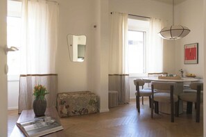 Un soggiorno con un divano, un tavolino da caffè con un libro e un vaso, una zona pranzo con tavolo e sedie, e un'ampia finestra con tende.
