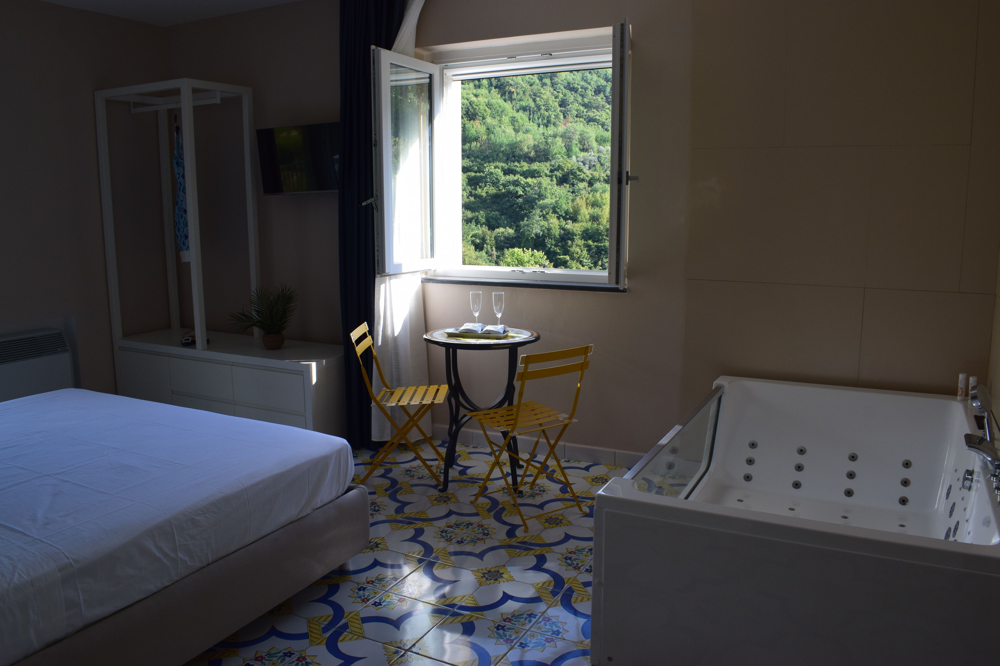 Una camera d'albergo con un letto, un tavolino con due bicchieri, una poltrona, una vasca da bagno e una finestra vista sul verde.