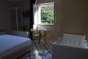 Una camera d'albergo con un letto, un tavolino con due bicchieri, una poltrona, una vasca da bagno e una finestra vista sul verde.