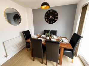A dining room with a wooden table set for four, black chairs, a round wall clock, and a radiator. - Prime Location Modern Home, Sleeps 5, Free Parking (Coventry)