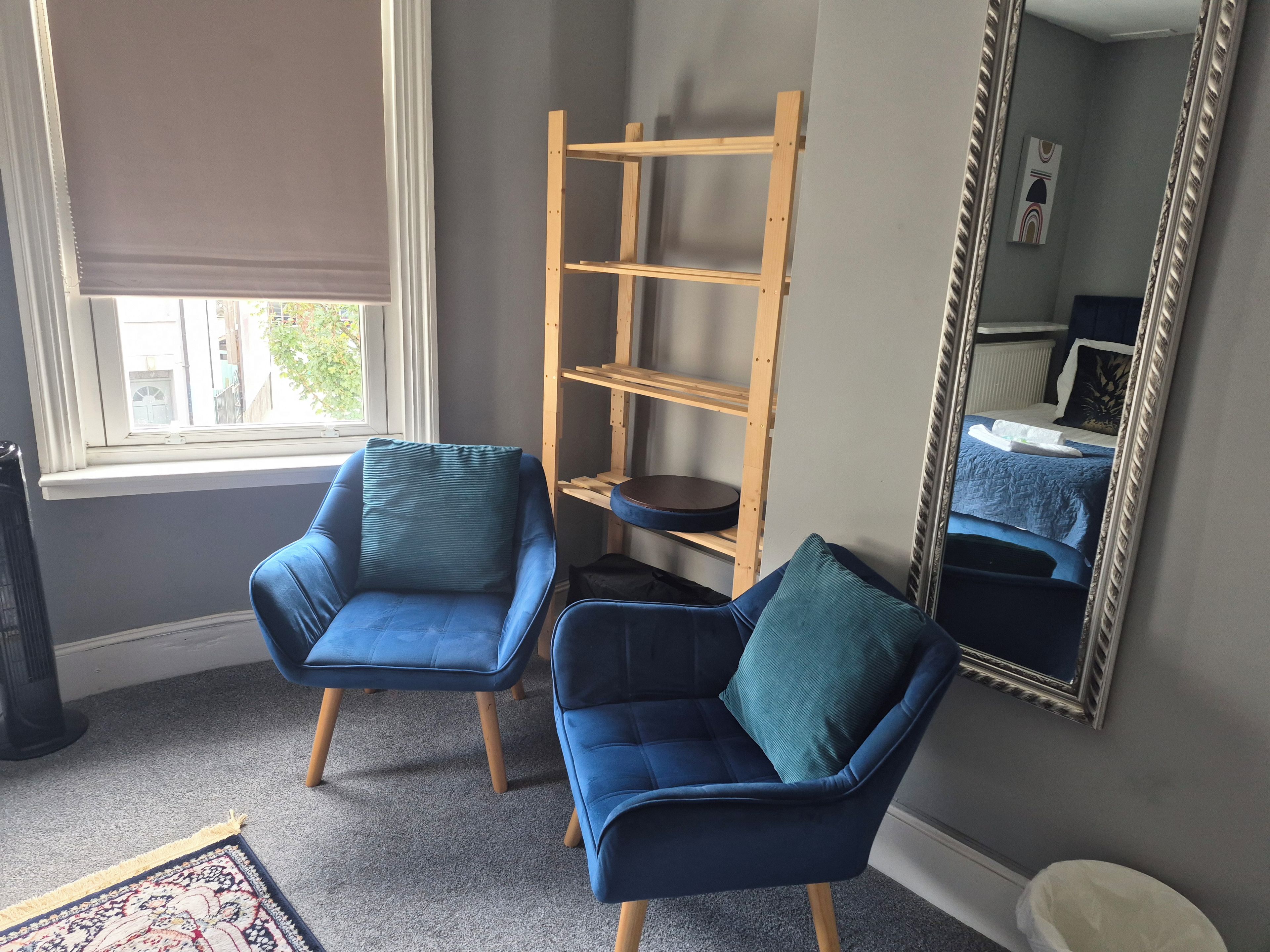 A room with two blue armchairs, a wooden shelf unit, a large mirror, and a window with blinds.