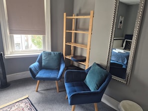 A room with two blue armchairs, a wooden shelf unit, a large mirror, and a window with blinds.