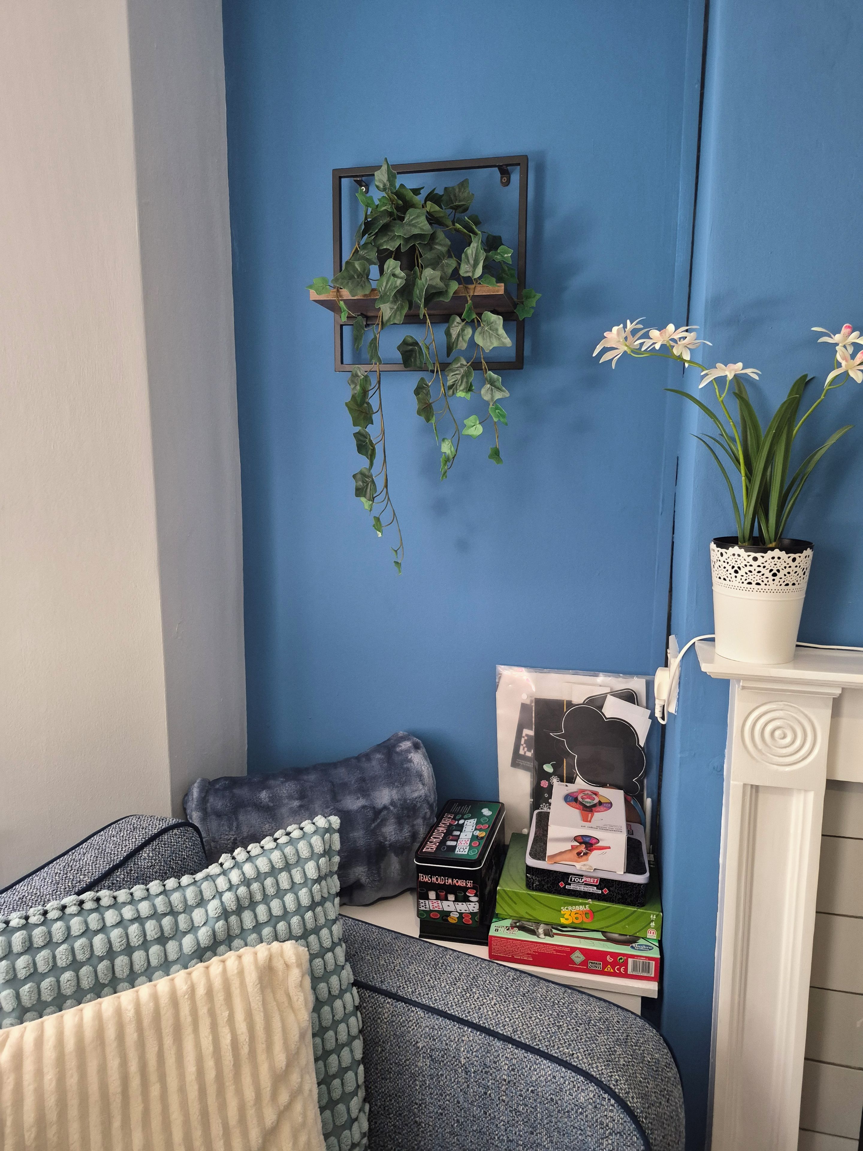 A corner of a room with a blue wall, a hanging plant, a white fireplace, and a shelf with books and a plant.