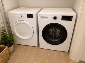 Two front-loading washing machines in a laundry room with a potted plant and a wicker basket. - Arnona Apartment - New & Modern WIth Private Mamad (Jerusalem)