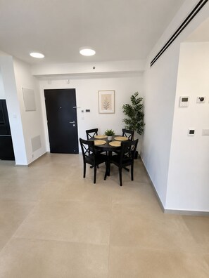 A room with a dining table and chairs, a black door, a small plant, and a thermostat on the wall. - Arnona Apartment - New & Modern WIth Private Mamad (Jerusalem)