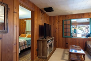 A wooden room with a bed, a television on a stand, a window with a view of the outdoors, and a small table with books. - Cozy log cabin on George Lake in Rhinelander, Wisconsin. (Rhinelander)