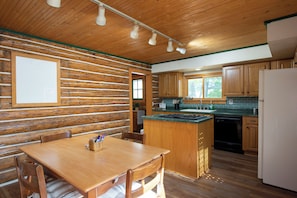 A cabin interior with a wooden dining table, kitchen area with wooden cabinets, and a built-in island with a stove. - Cozy log cabin on George Lake in Rhinelander, Wisconsin. (Rhinelander)