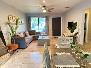 A modern living room with a sofa, coffee table, and dining table. There is a ceiling fan, a large window, and a door leading outside. - Modern 4-bedroom Oasis in North Miami w/ HOT TUB (Miami)