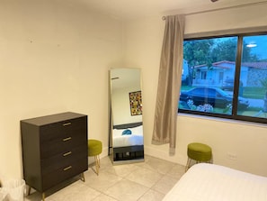A bedroom with a bed, a dresser, a mirror, a window with a view of a house, and a green stool. - Modern 4-bedroom Oasis in North Miami w/ HOT TUB (Miami)