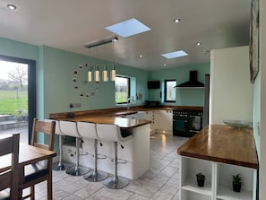 A modern kitchen with a central island, bar stools, and a dining area with a wooden table and chairs. - Woodland Forge - The Stables (Over Whitacre)