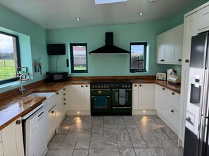 A kitchen with a central stove, white cabinets, and a window overlooking a garden. - Woodland Forge - The Stables (Over Whitacre)