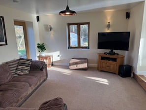 A living room with a sofa, ottoman, TV stand, and a large window. - Woodland Forge - The Stables (Over Whitacre)