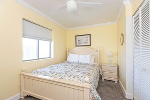Une chambre à coucher comprenant un lit, une table de chevet, une lampe, une fenêtre avec des stores, un ventilateur de plafond et une porte avec un miroir.