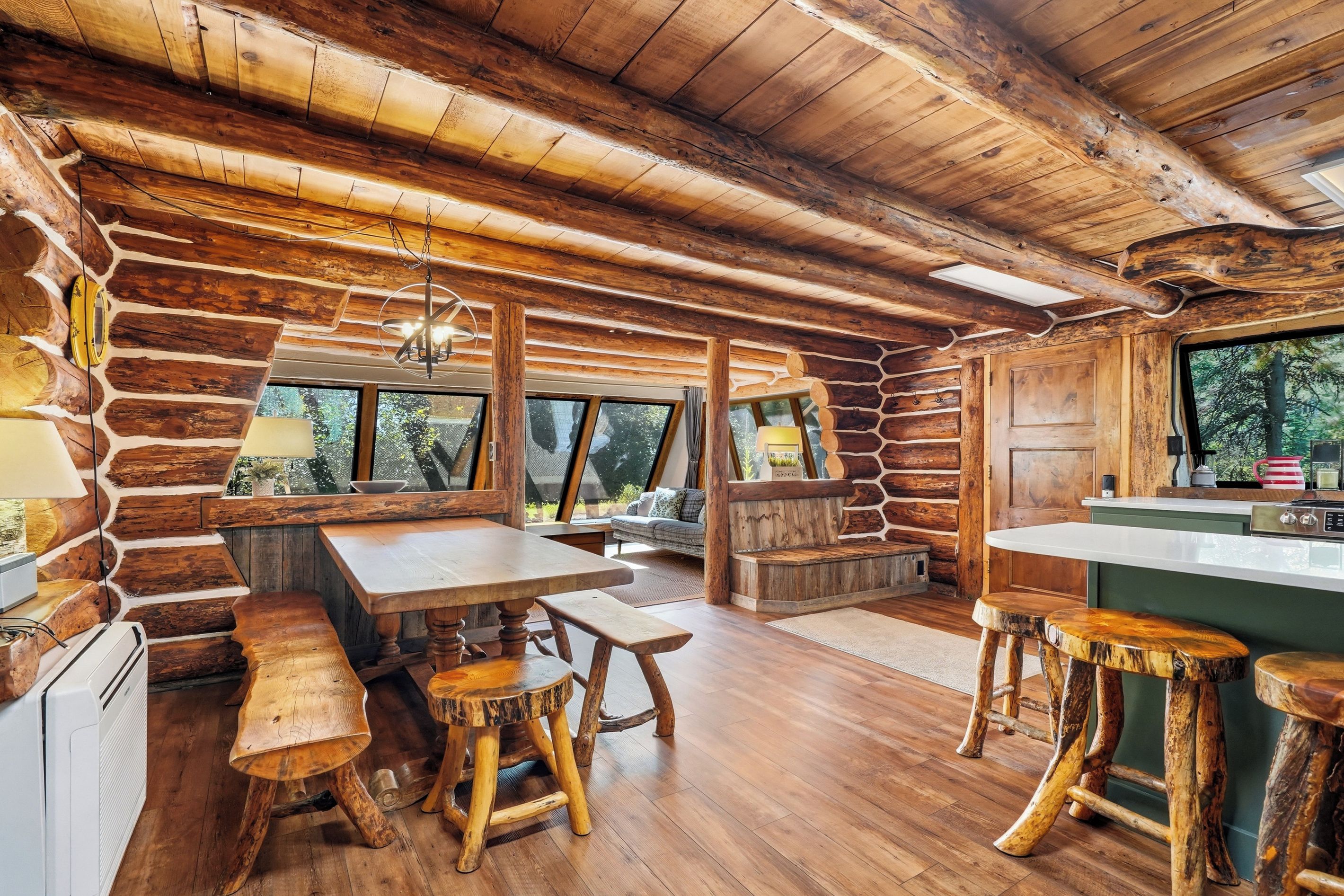 Interior rústico de una cabaña con una mesa de comedor de madera, bancos, una zona de cocina y ventanas amplias que ofrecen vistas al exterior.