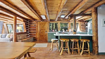 Interior rústico de una cabaña con techo de madera, zona de comedor con mesa y bancos, cocina con barra y un ventanal con vistas a la vegetación.