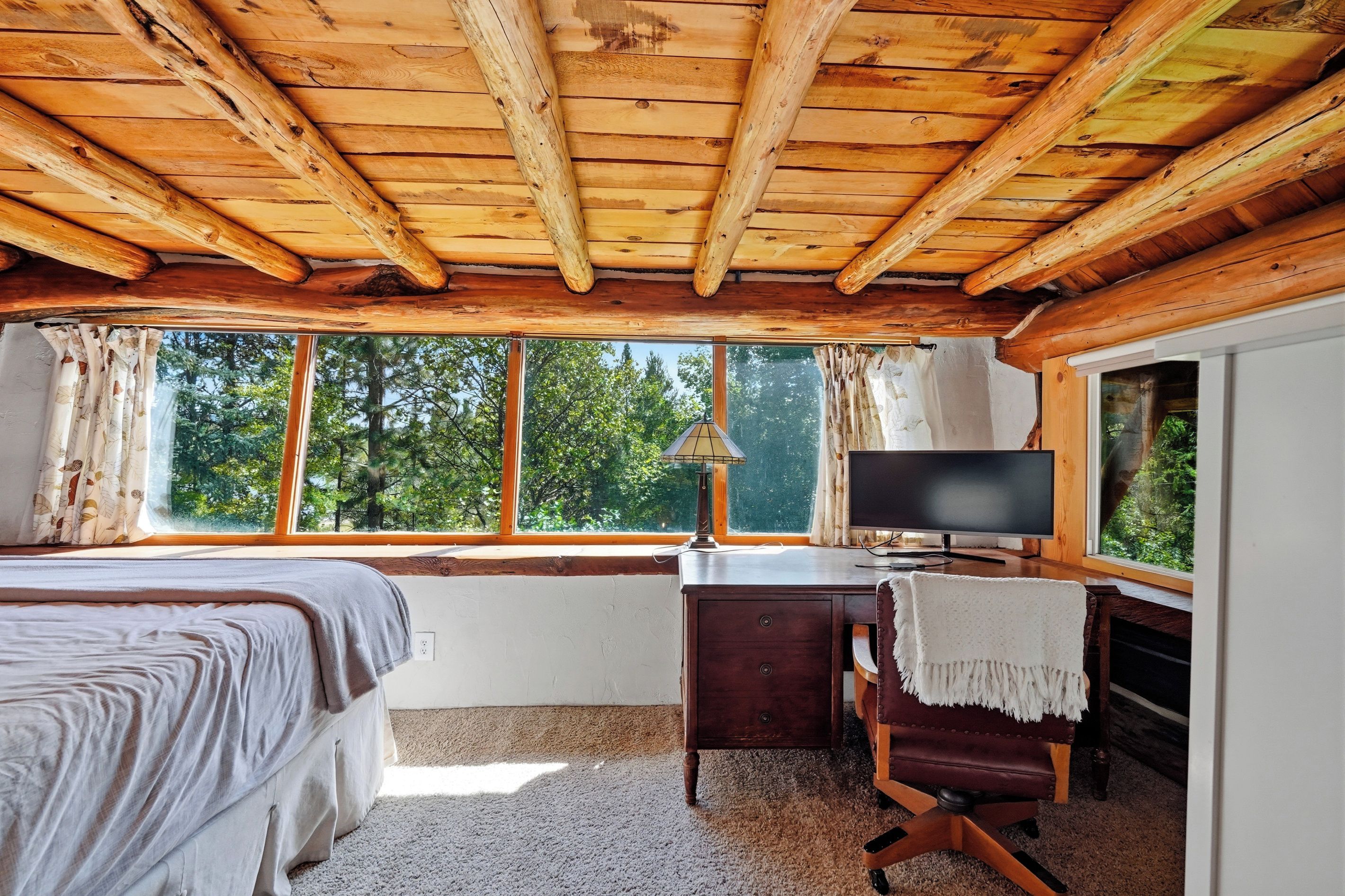 Un dormitorio con techo de madera, una cama, un escritorio con lámpara, un televisor y vistas a árboles a través de ventanas amplias.