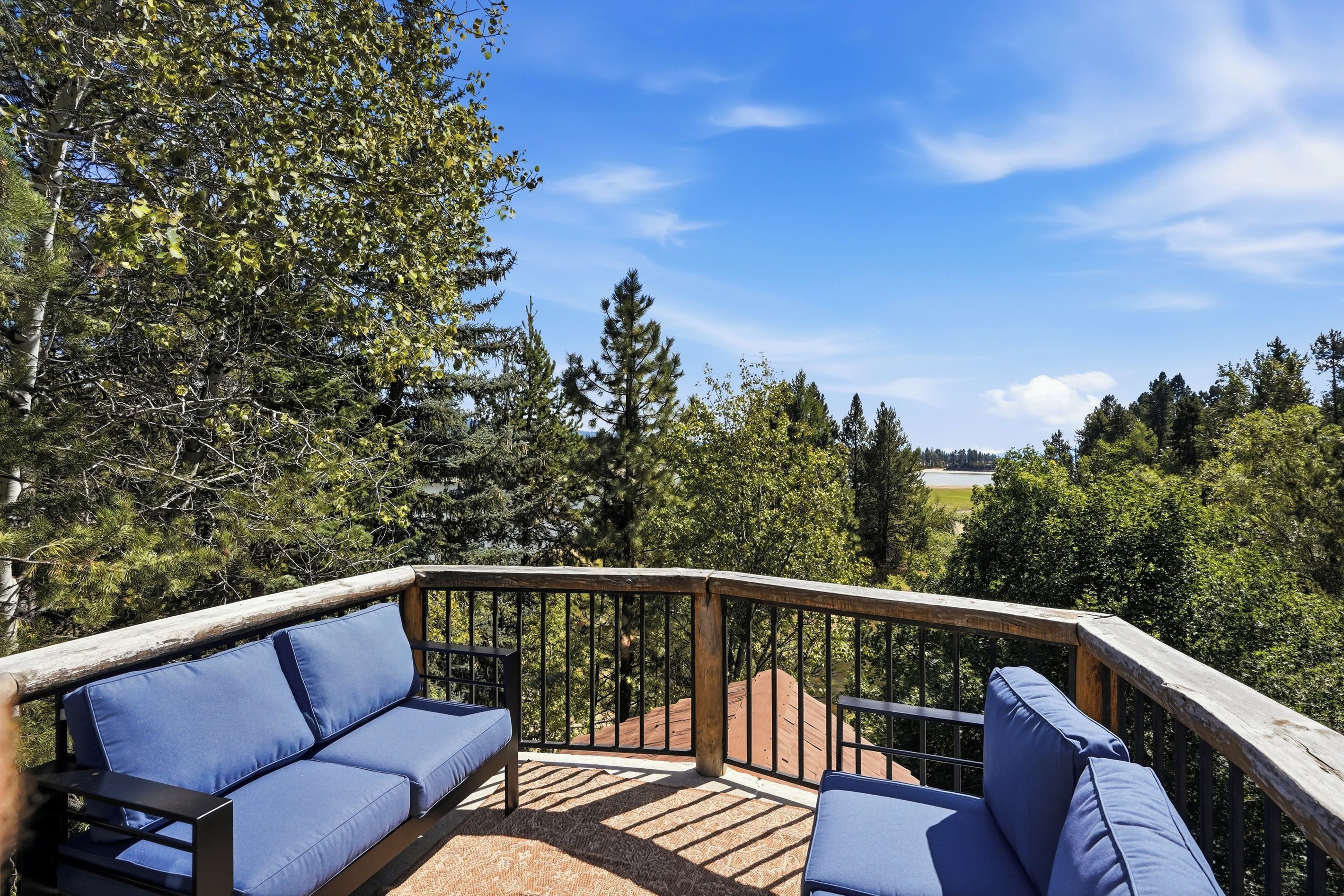 A deck with a blue cushioned seating area, surrounded by a wooden railing, overlooking a forested landscape.