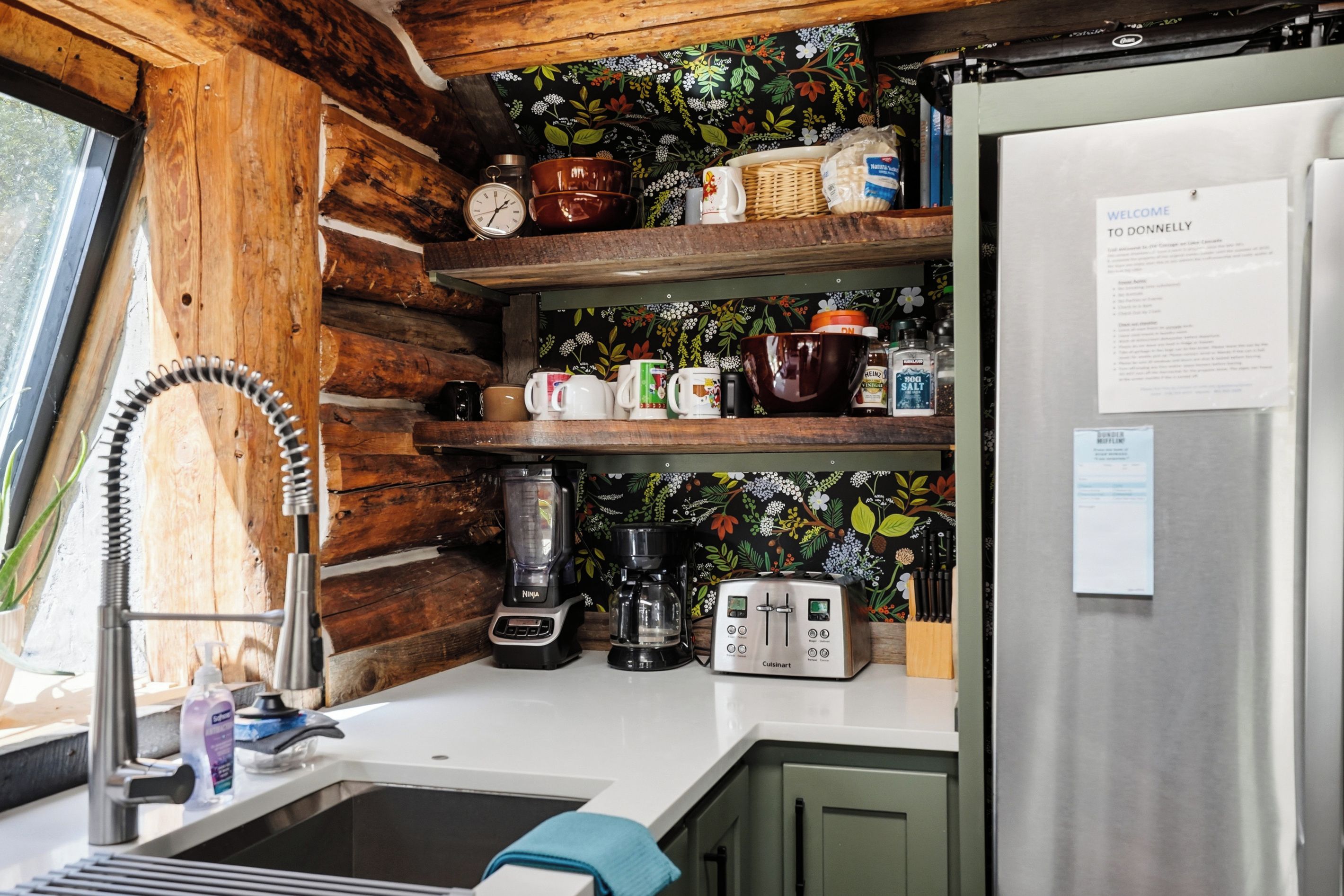 A compact kitchen with wooden shelves, a coffee maker, a blender, and a refrigerator.