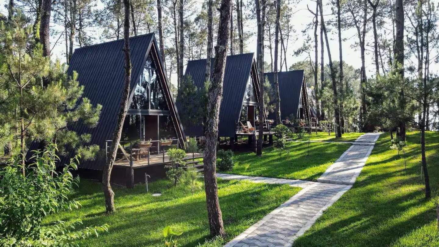 A row of cabins with sloped roofs, surrounded by trees and a paved walkway.