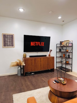 A living room with a flat-screen TV mounted on the wall, a wooden cabinet, a round coffee table, and a metal shelving unit with books and decorative items. - Furnished Extended Stay Near Downtown Indy (Indianapolis)