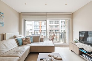 A modern living room with a sofa, a coffee table, and a television. There is a balcony with a view of the cityscape. - Lectus II 0101 met garage (Nieuwpoort)