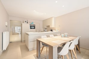 A modern kitchen with a central island, white cabinetry, and a dining area with a wooden table and chairs. - Lectus II 0101 met garage (Nieuwpoort)