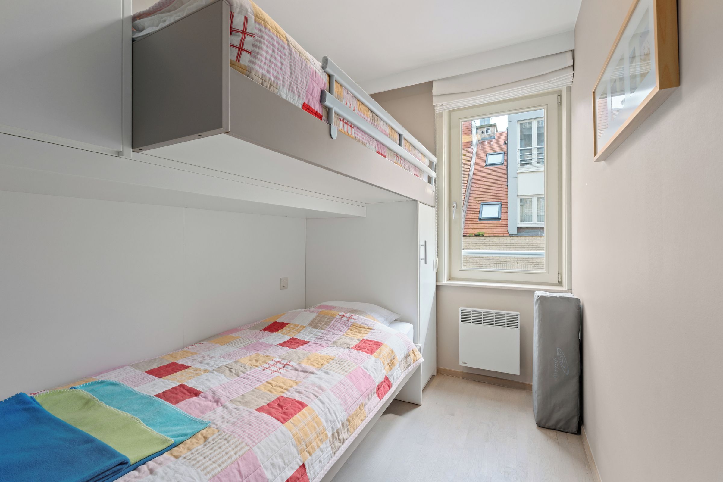 A small, single-bed room with a bunk bed above, a window with a view of buildings, and a radiator.