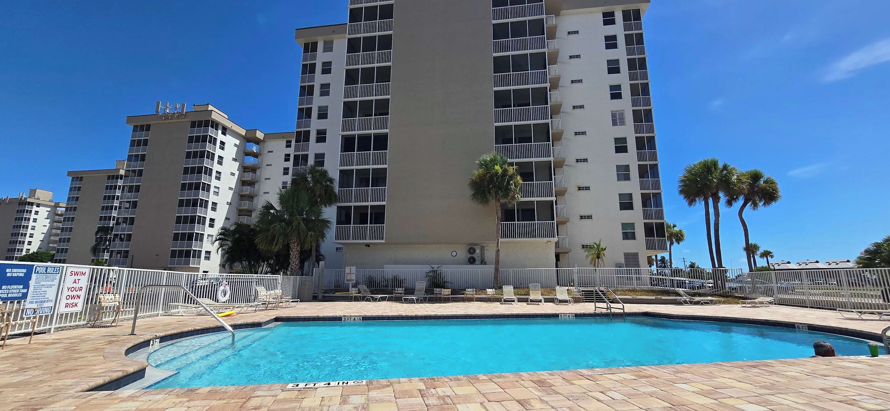 A swimming pool with a 3 ft 4 in depth, surrounded by a fence, with palm trees and a multi-story building in the background.