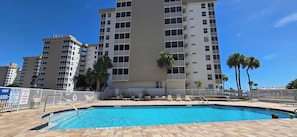 Piscina con una profundidad de 1 metro 20 centímetros, rodeada por una valla, con palmeras y un edificio de varios pisos al fondo.