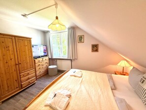 A bedroom with a bed, a wooden wardrobe, a window with curtains, a radiator, and a television displaying an advertisement. - Haus Heike - Holiday Apartment 1st Floor (Utersum)