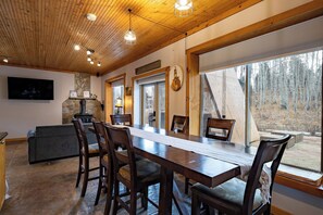 A wooden interior with a stone fireplace, a dining table with chairs, and a large window offering a view of a forest. - Mountain Retreat 5BR • Hot Tub • 25 Min to PC (Coalville)