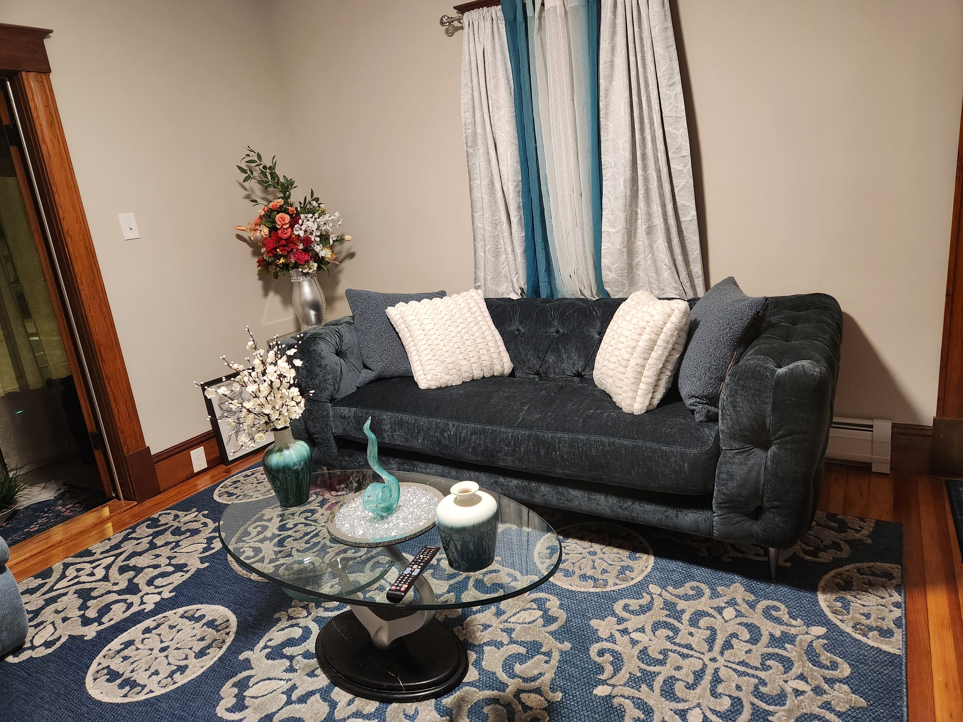 A living room with a dark blue sofa, white cushions, a glass coffee table with decorative items, a blue patterned rug, and a vase with flowers.