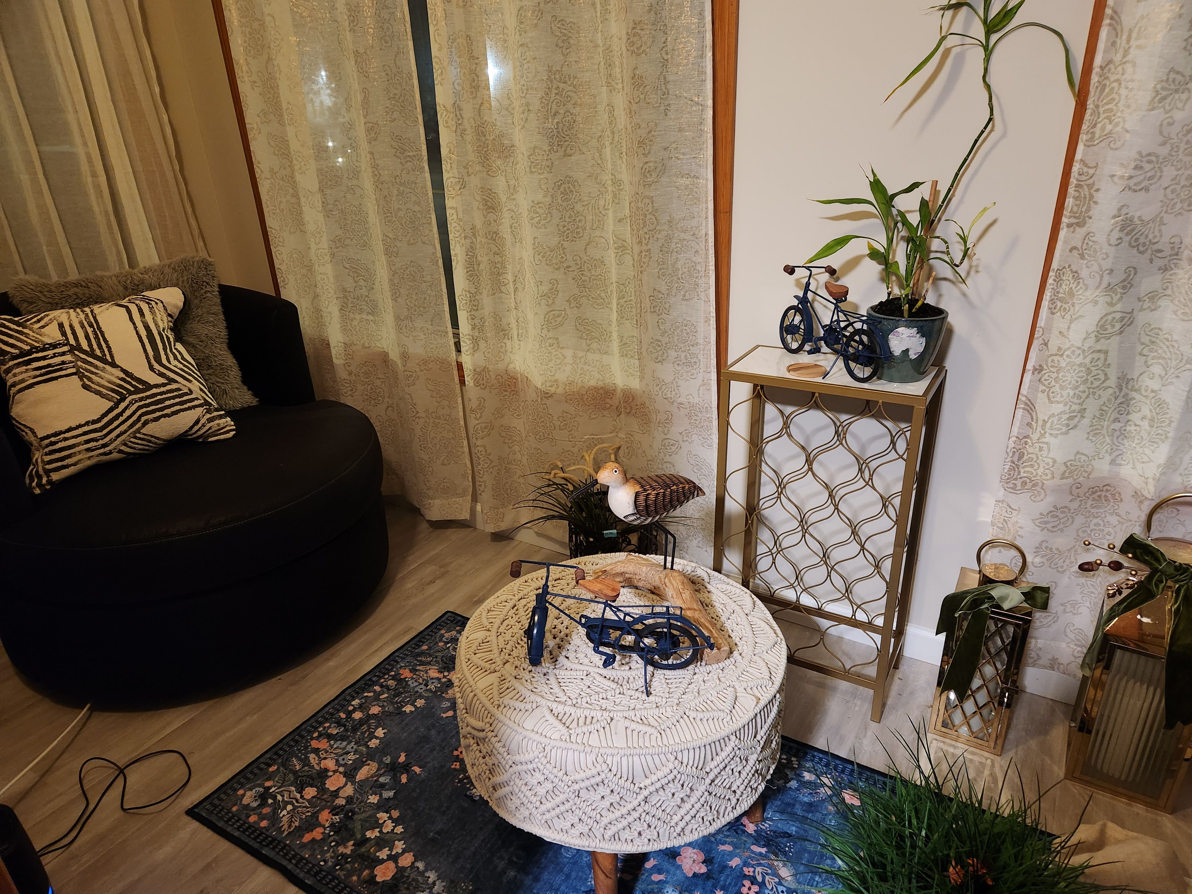 A living room with a black sofa, patterned cushions, a round woven ottoman, a small table with a plant, a decorative bird, and a bicycle model.