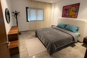 Ein Schlafzimmer mit Bett, Nachttisch, Kommode, Kleiderständer und einem Fenster mit Jalousien.