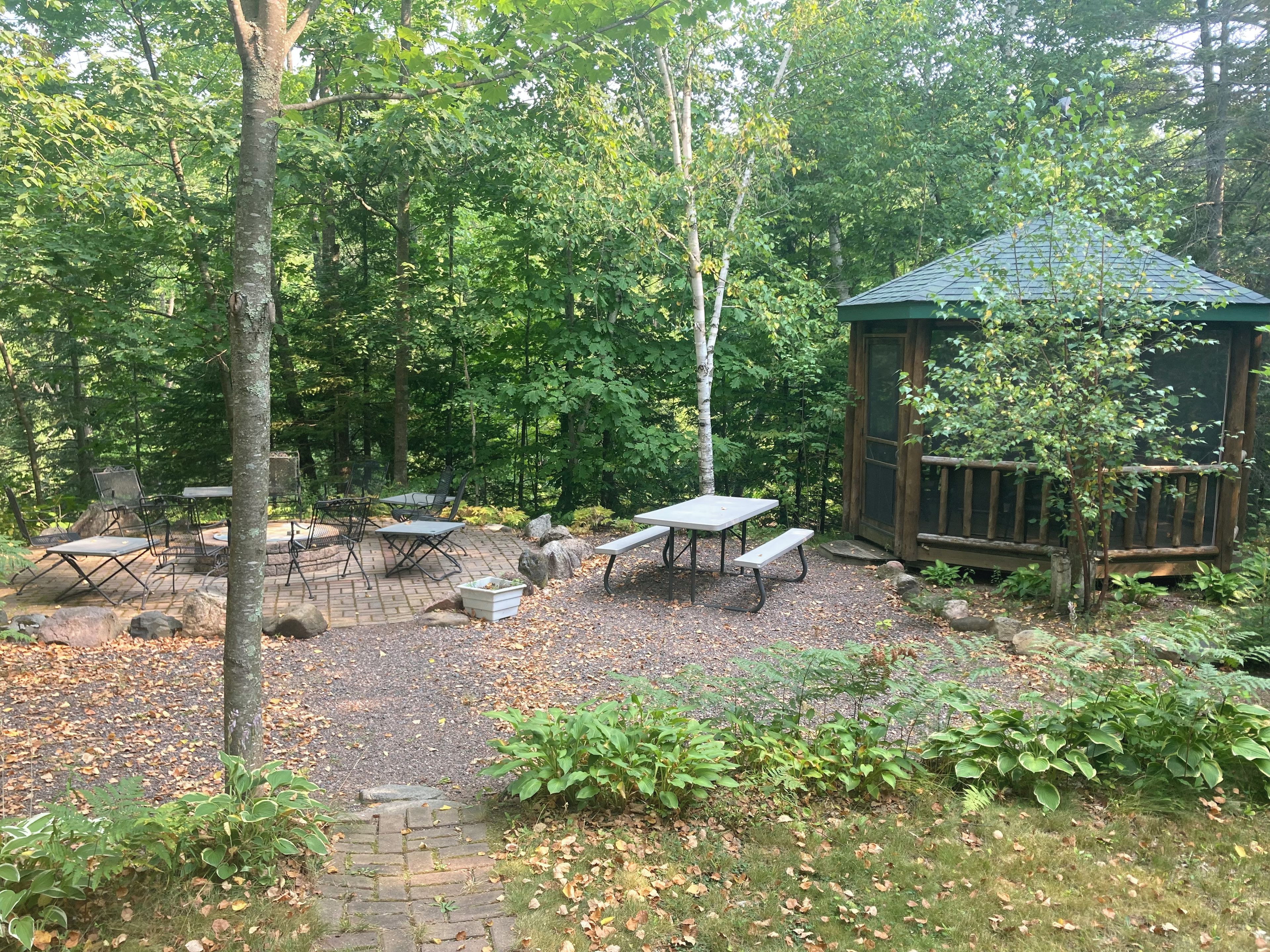 An outdoor patio area with picnic tables and chairs, surrounded by trees and a wooden structure.