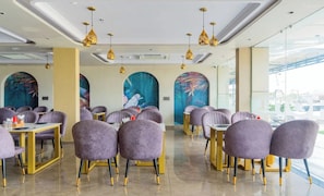 Een restaurant met paarse stoelen en gouden accenten, met grote ramen en kunst aan de muren.