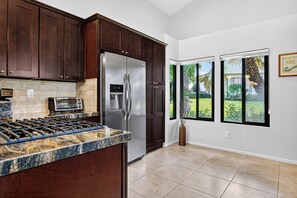 A kitchen with dark wood cabinets, a stainless steel refrigerator, a built-in oven, and a gas stove. There are two windows with a view of a garden. - 3-Bedroom Desert Oasis Just Steps to La Quinta Resort! (La Quinta)